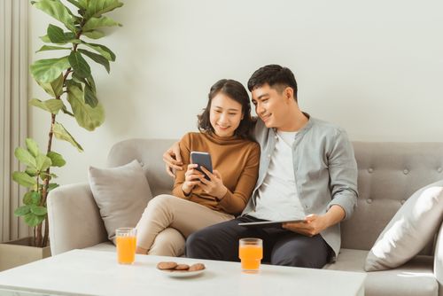 A smiling couple sitting on a couch in a modern living room, looking at something on one of their phones.