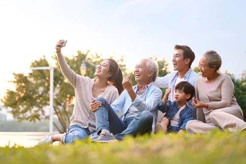 A family of all ages sitting on a grassy hill smiling while taking a selfie together.