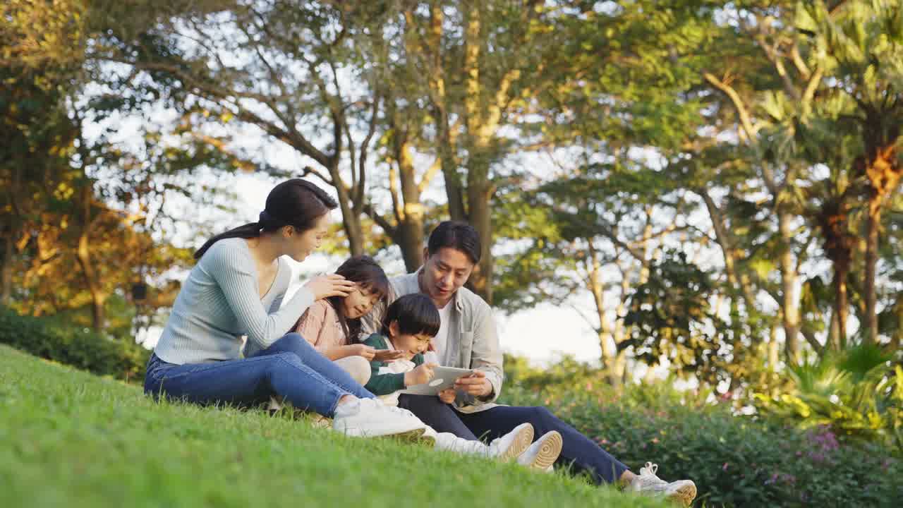 A young family sitting together on a grassy hill watching a video on a tablet computer.