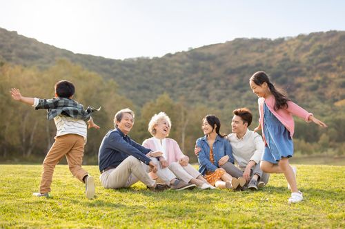 Two children laughing and running around their parents and grandparents who are sitting and watching the children in a wide grassy field.