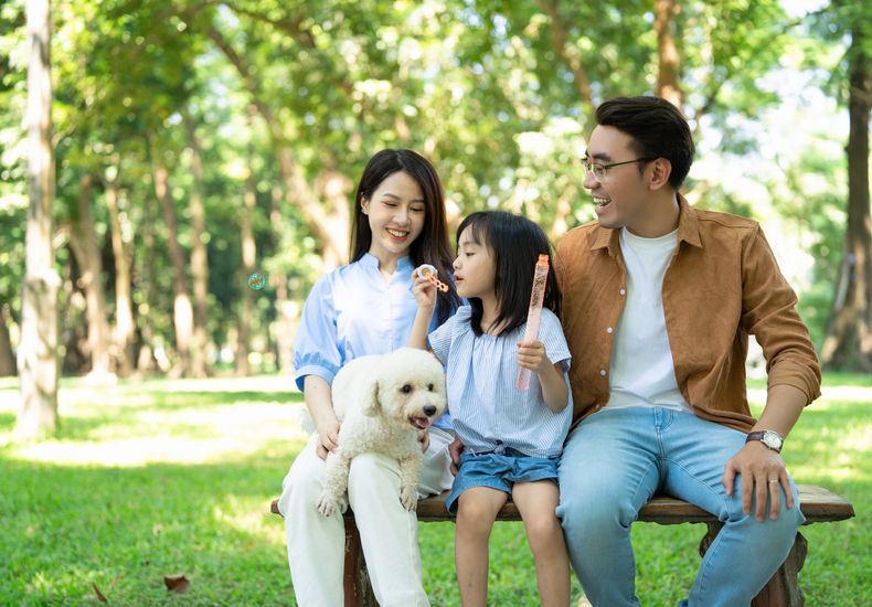 A family with their dog sitting on a bench in a lush park. The child is blowing bubbles while their smiling parents watch.