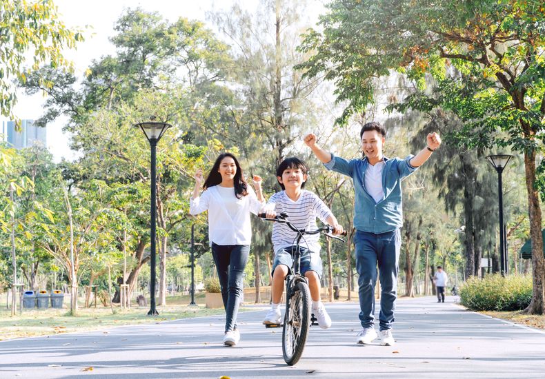 A child smiling while riding a bicycle down a calm, tree-lined path while their parents run behind encouraging their child.